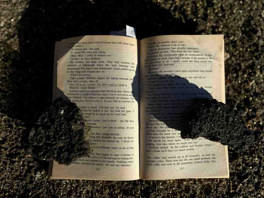 Open book on gritty floor held down with volcanic rocks, showing murder mystery plot annotated with underlining : 'Hart Fell is all I needed. You didn't need to climb it' and 'if Flip didn't need to climb Hart Fell, I wonder if she needed to do any travelling at all. I mean, could she have solved the puzzle from her bedroom?'