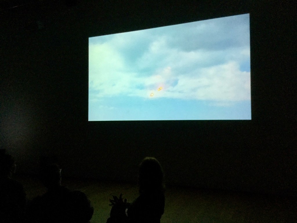 Morse code torchlight in a stormy sky, seen as a projected film in a dark gallery with audience