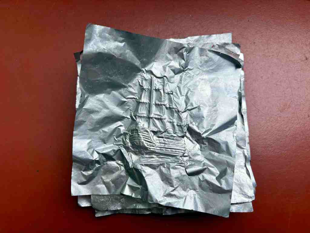 Stacked square flattened silver foil wrappers on a dark red painted surface, with imprinted rubbing of a tall ship with sails in the middle of top foil square